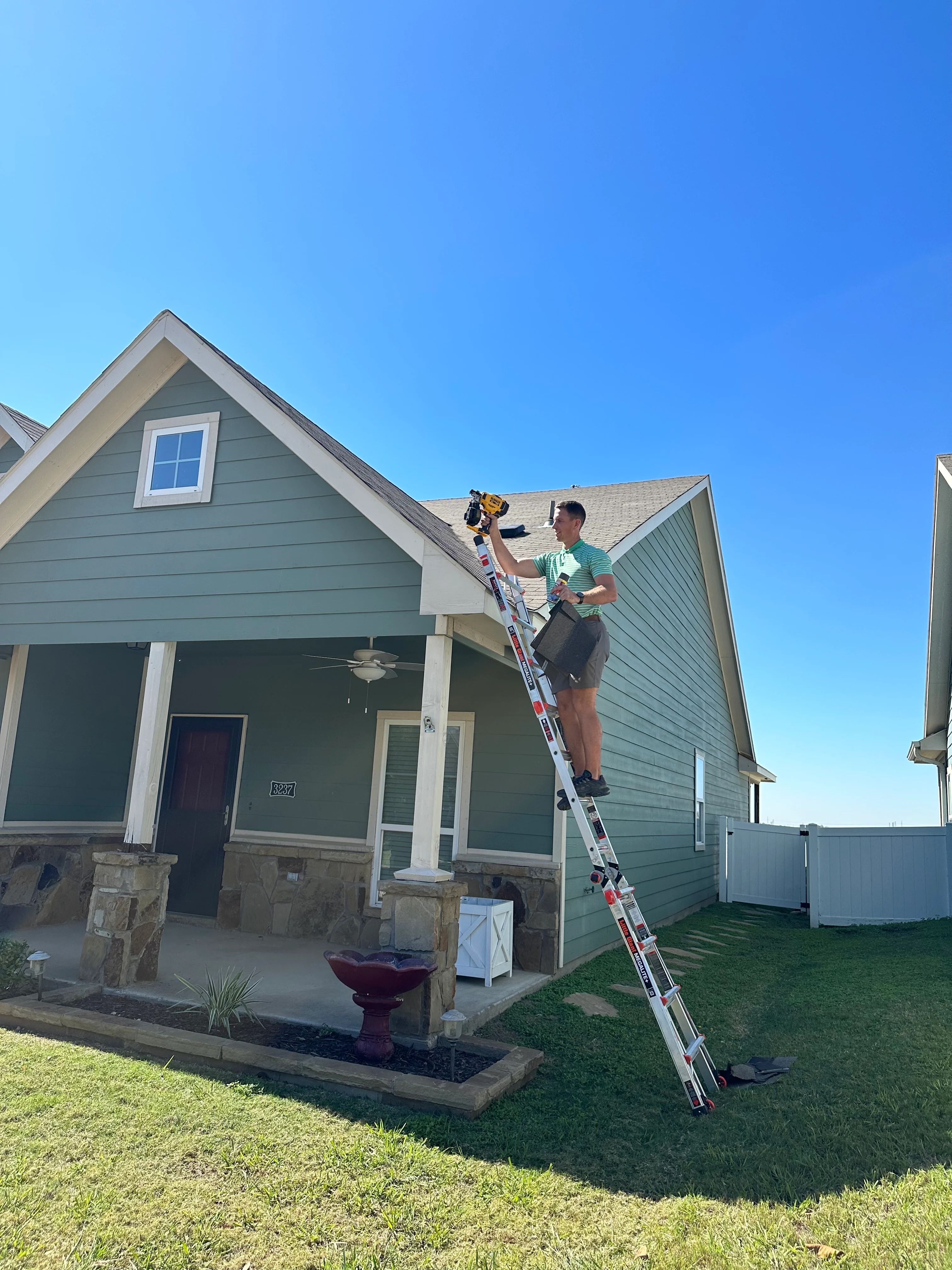 Technician on ladder inspecting gray shingle roof of residential home with stone foundation