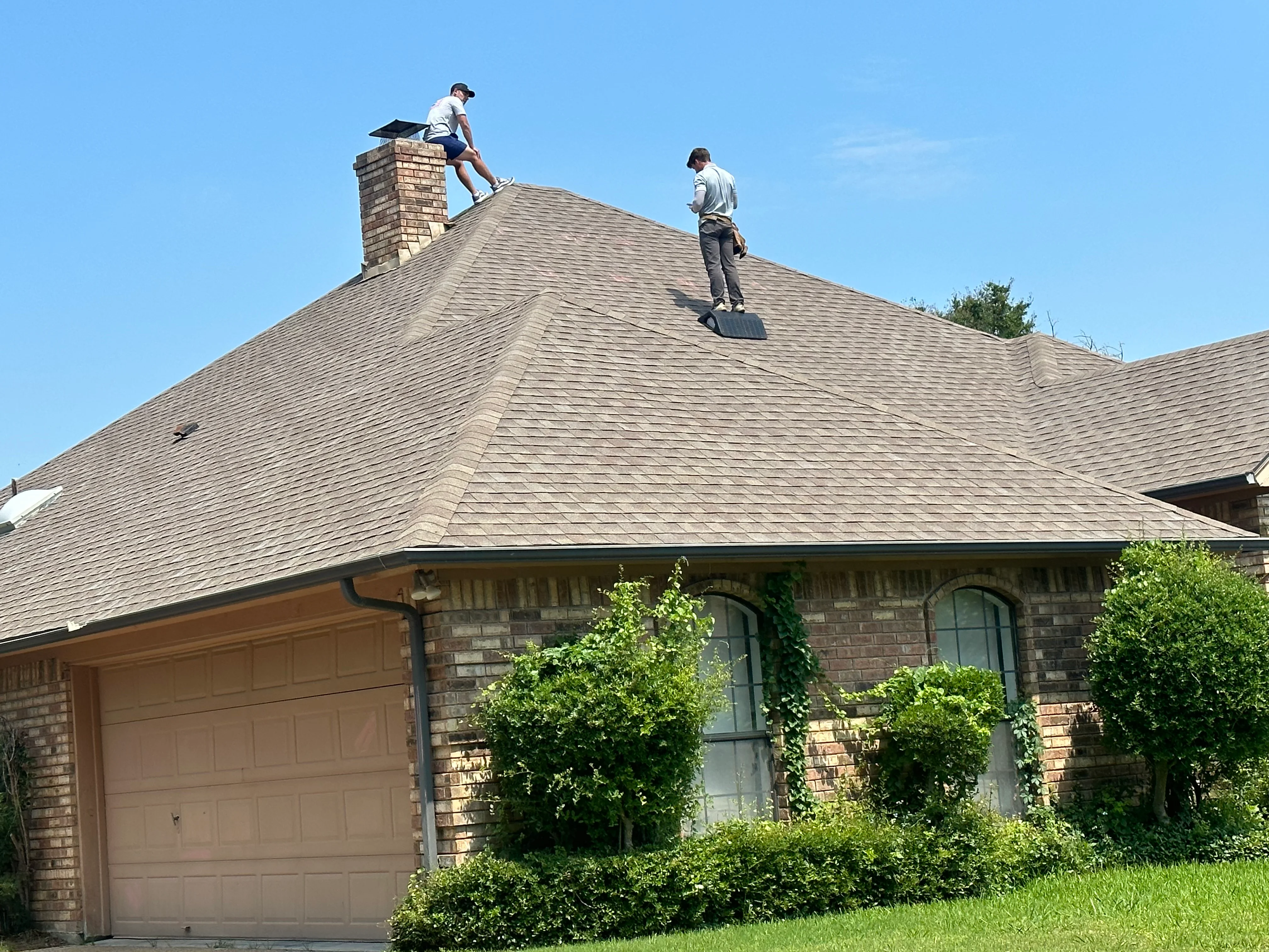 Two roofers installing gray architectural shingles on steep residential roof with brick chimney