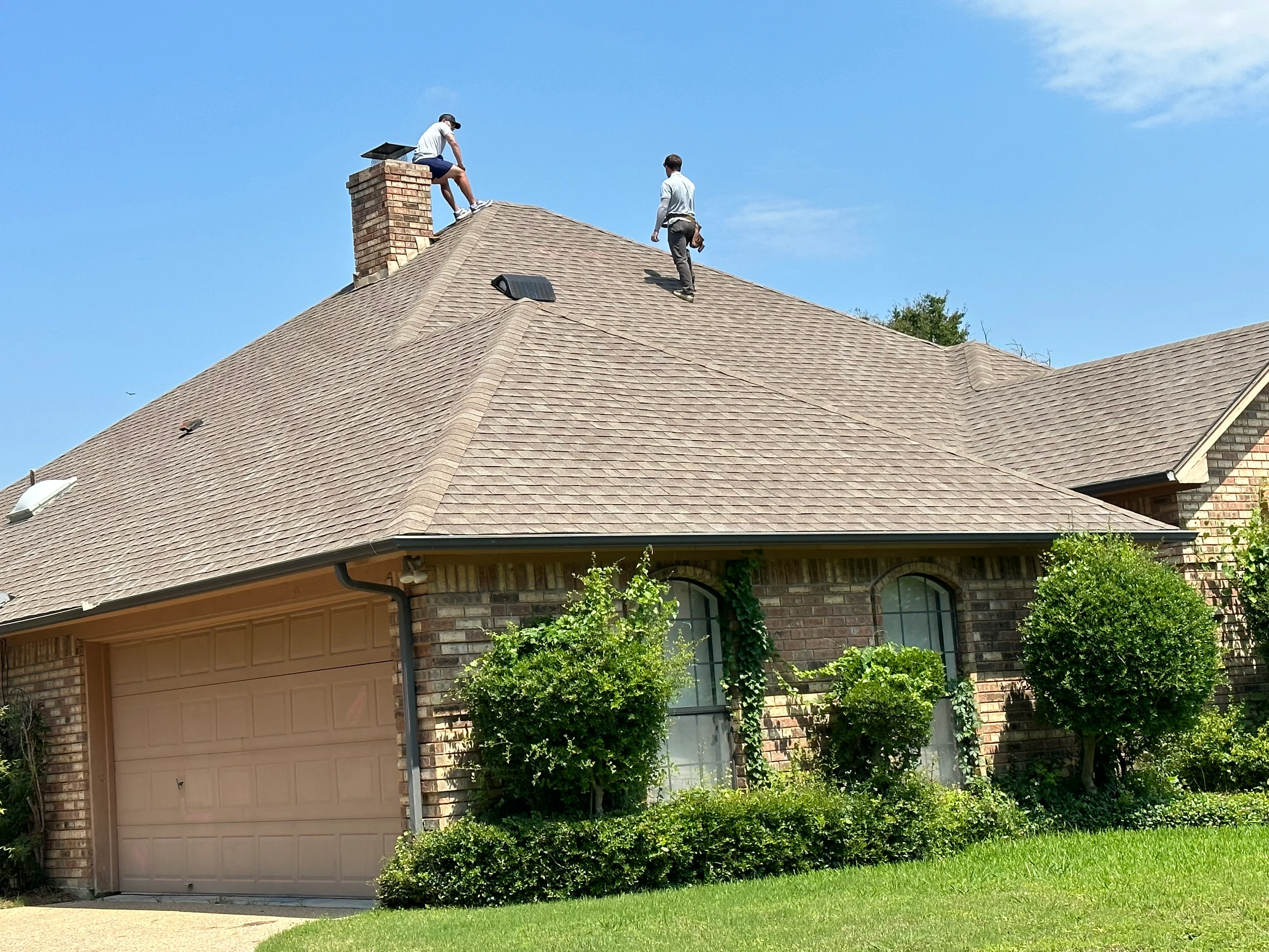 Roofers installing gray architectural shingles on pitched residential roof with brick chimney