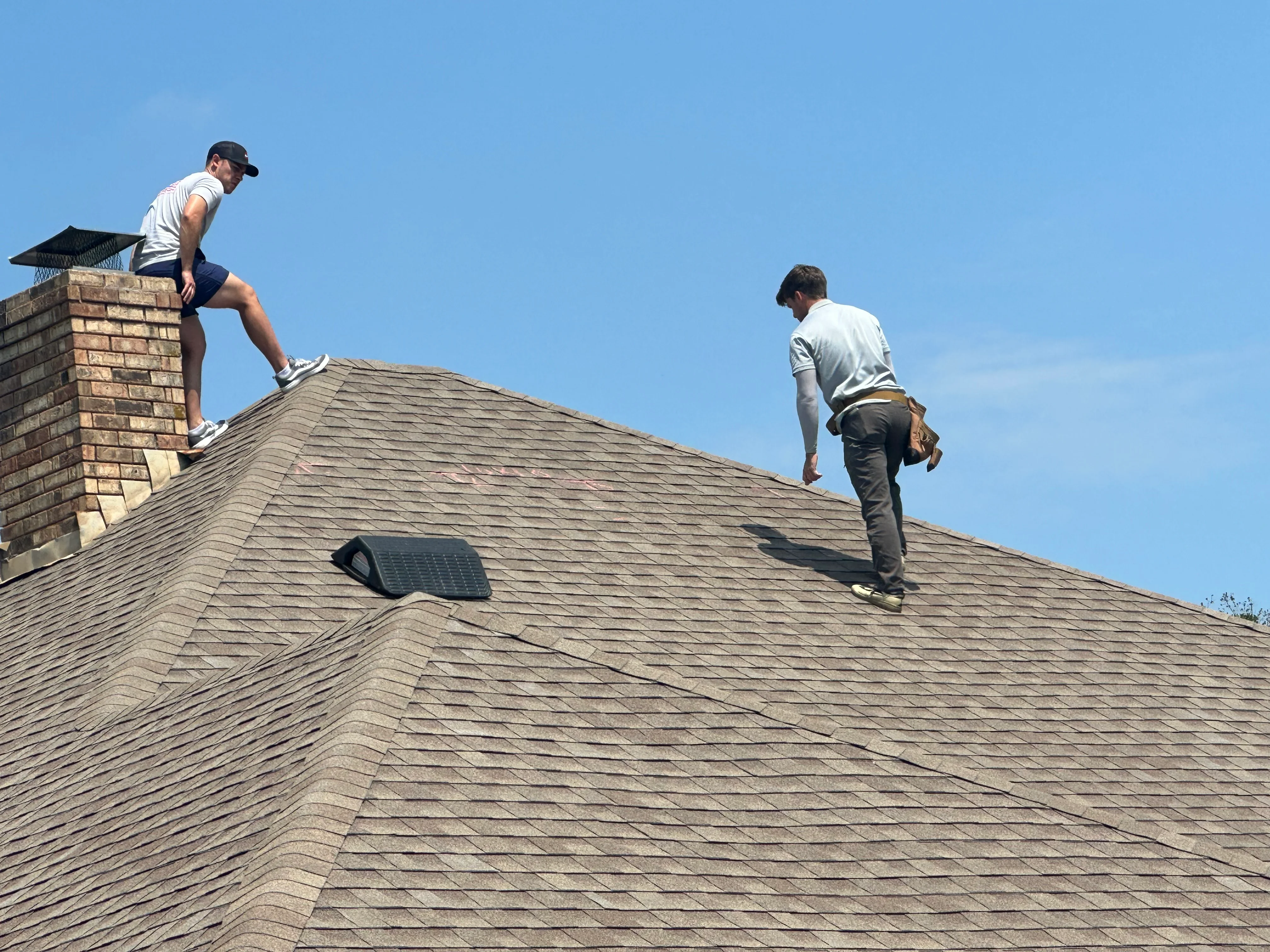 Roofers installing gray asphalt shingles on pitched residential roof with brick chimney