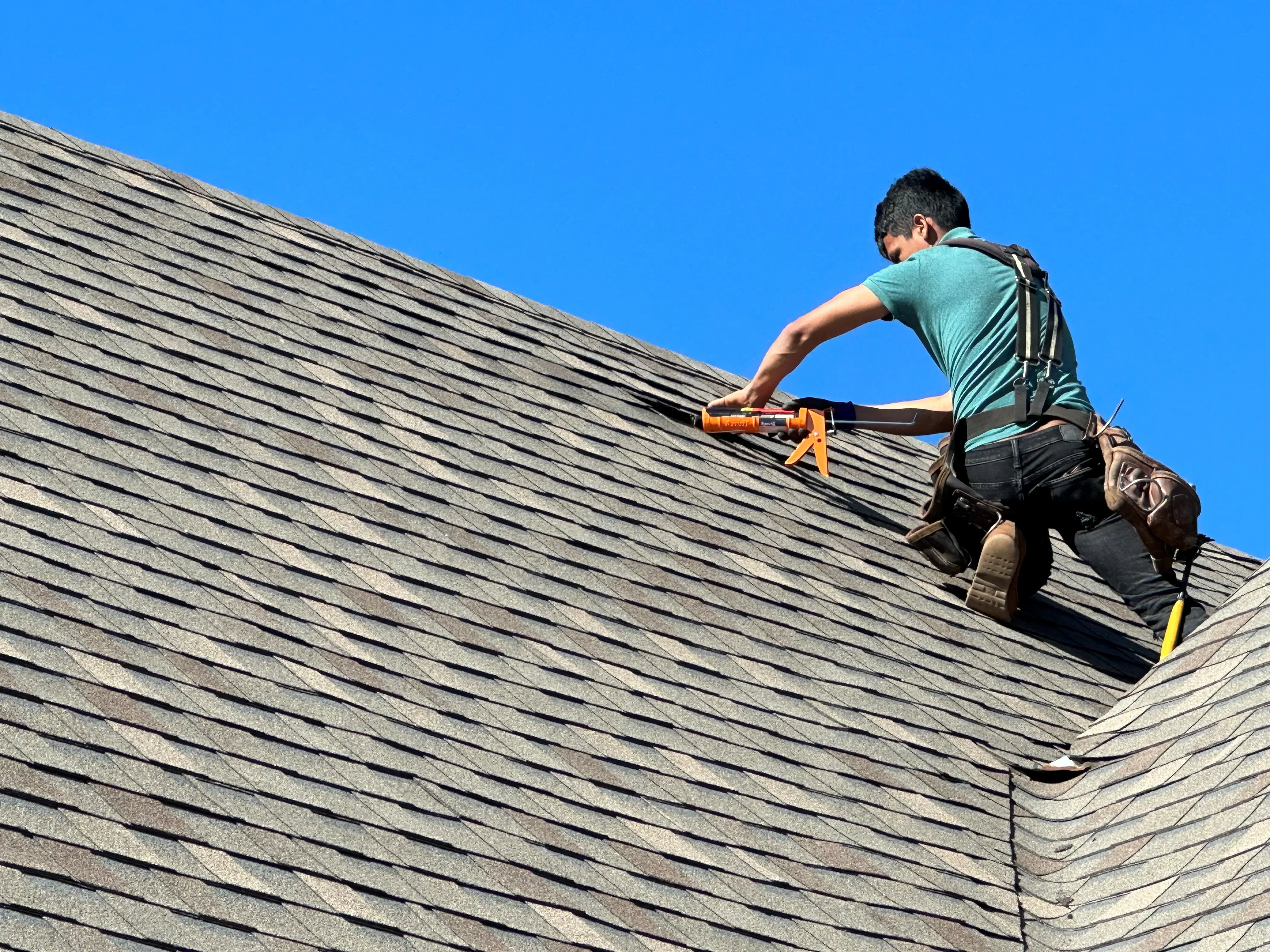 Roofer installing asphalt shingles on pitched residential roof with caulking gun