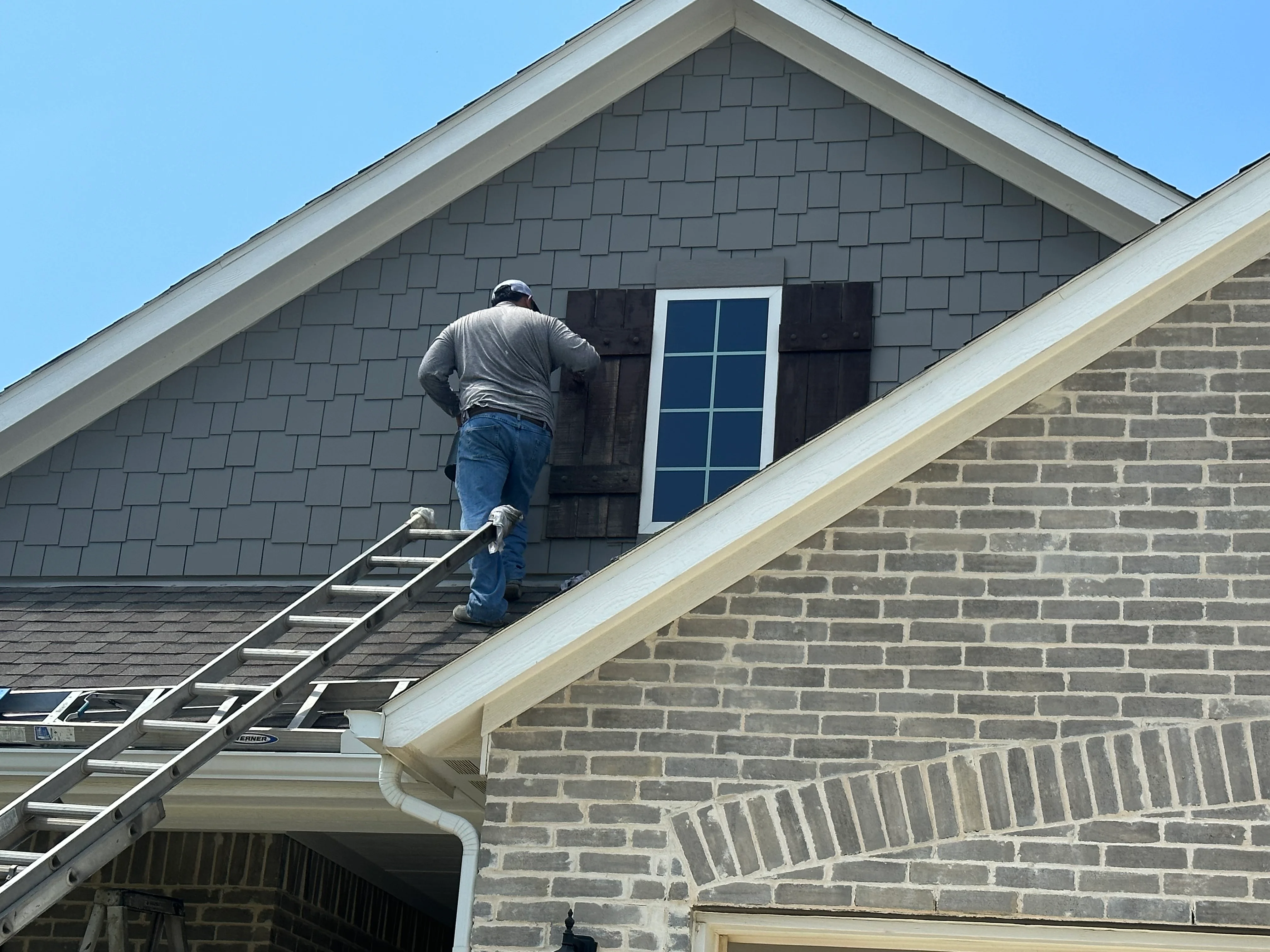 Worker installing gray architectural shingles on gable end with white trim and window flashing