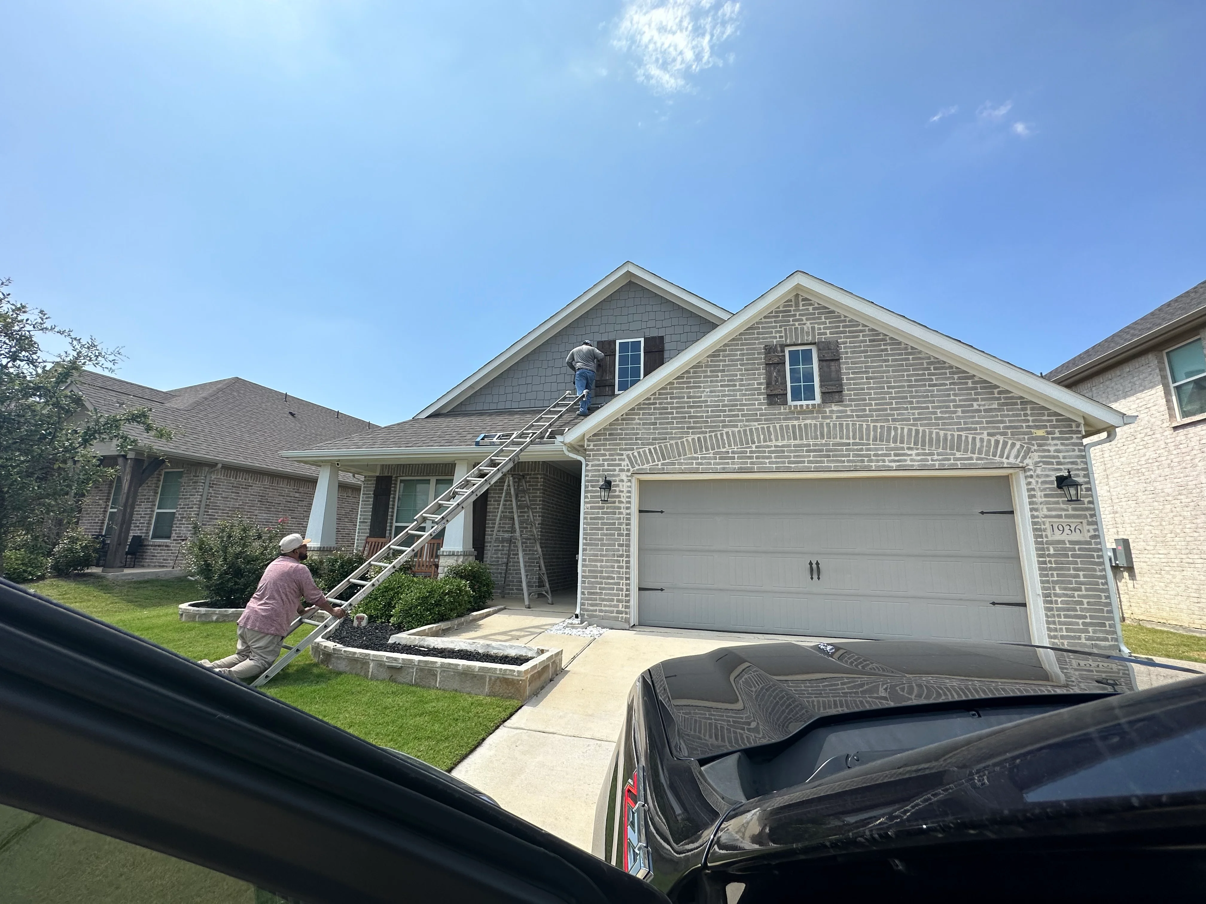 Roofer on ladder inspecting two-story brick home with asphalt shingle roof and gable vents