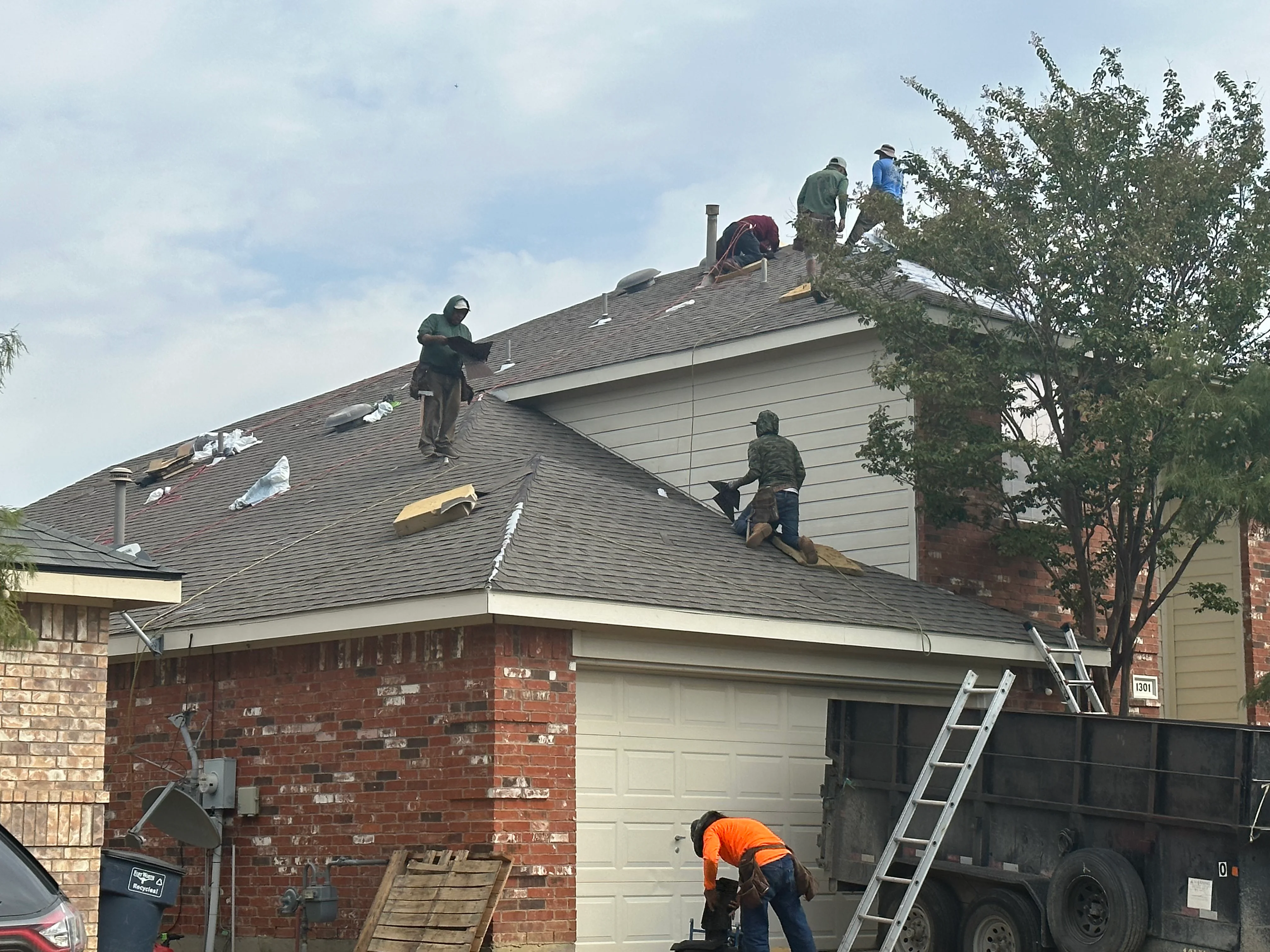 Roofing crew installing asphalt shingles on residential home with brick and siding exterior